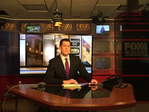 a man in a suit and tie sits at a Fox news anchor desk